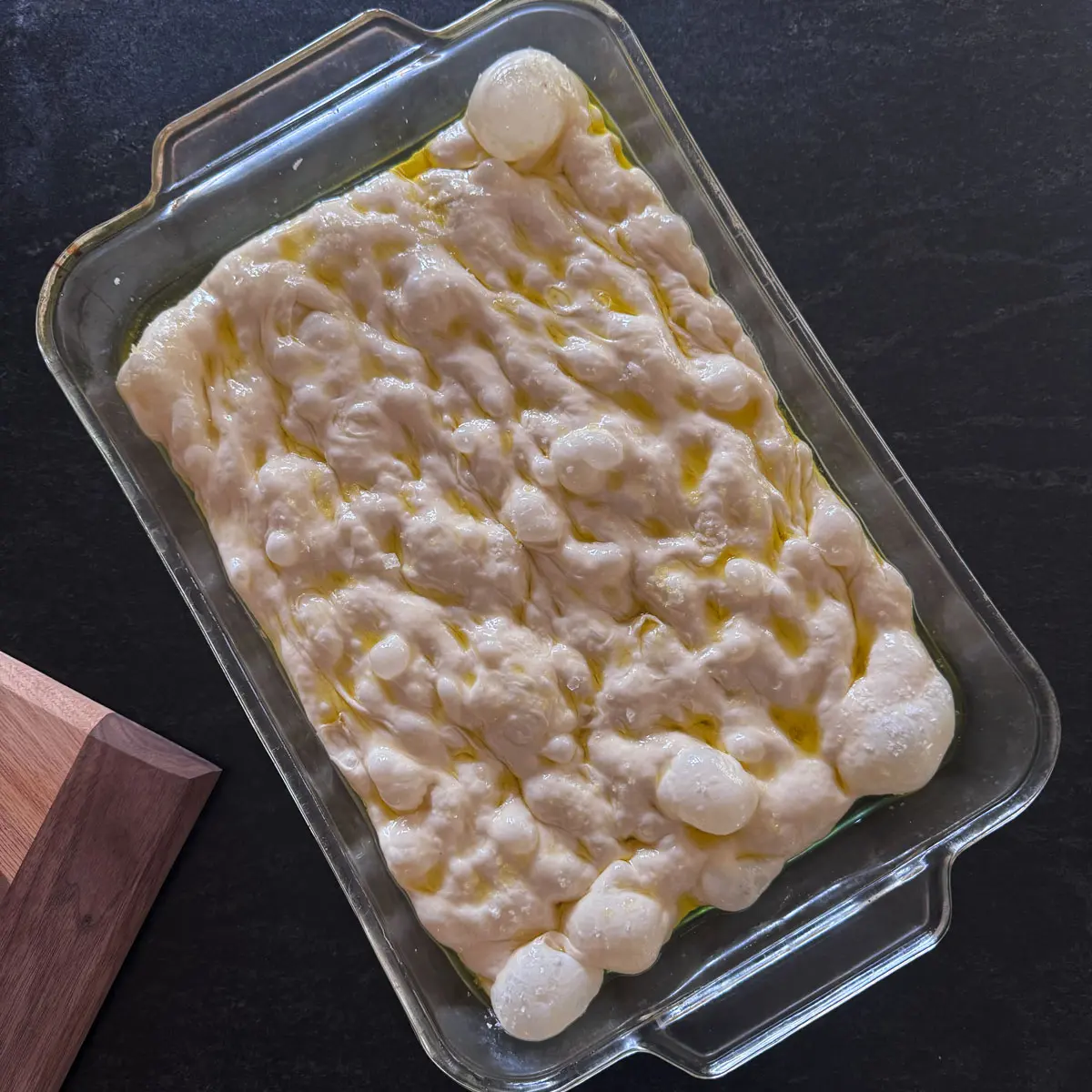 High-hydration focaccia dough proofing in an oiled glass 9×13 pan, its surface covered in large rounded bubbles and pooled olive oil.
