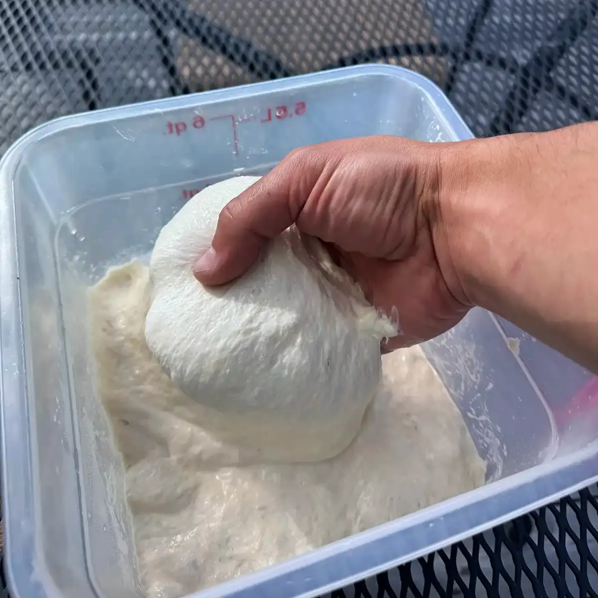 A hand lifts a soft, stretchy ball of pizza dough from a plastic container on an outdoor metal table.