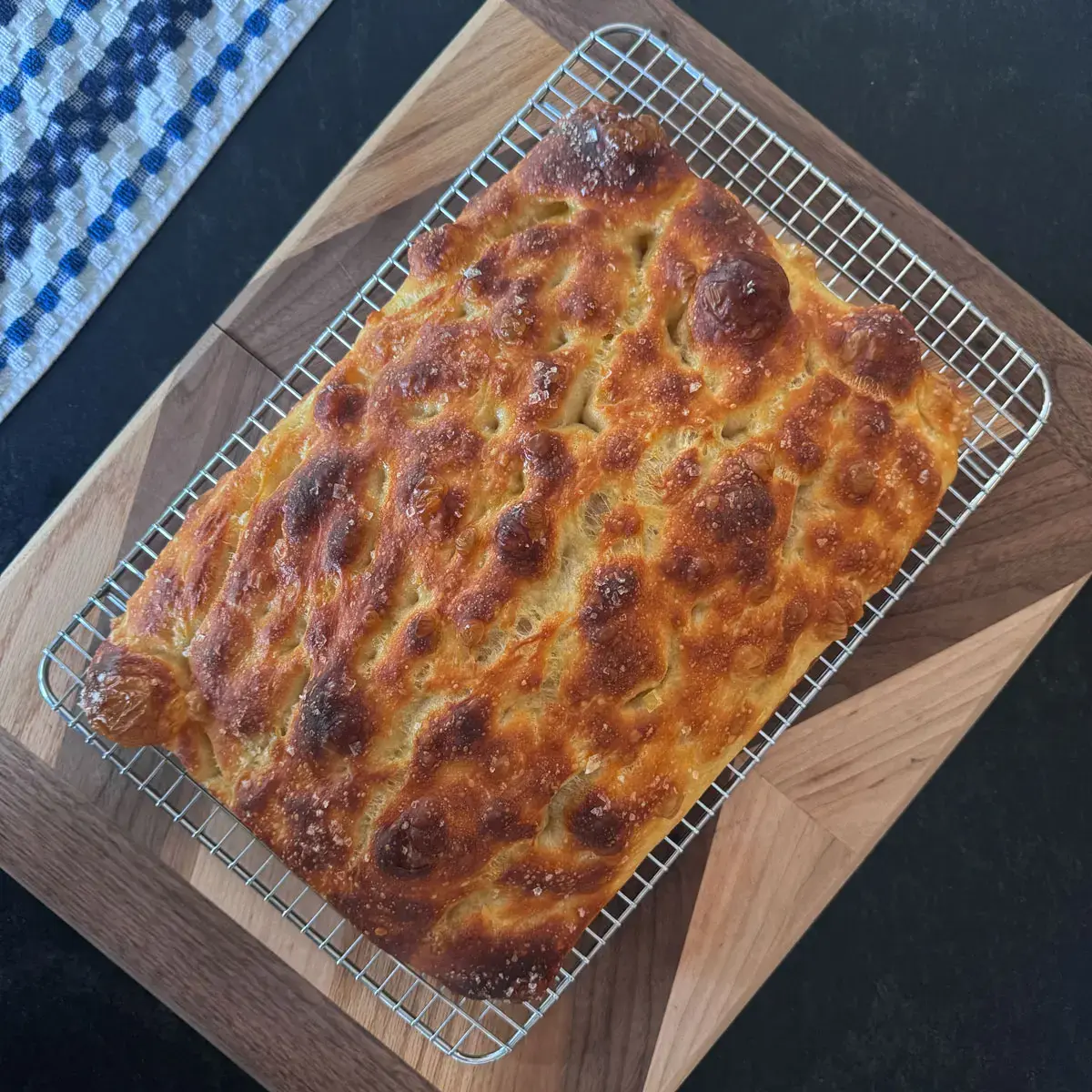 Golden-brown focaccia with a deeply dimpled, blistered top scattered with flaky salt, cooling on a wire rack over a wooden board.